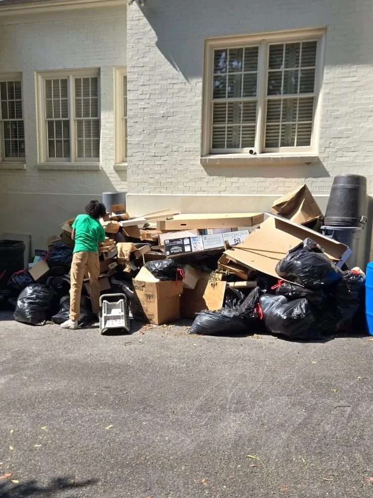 A crew member stacking flattened cardboard and bagged trash on the driveway beside a painted brick house