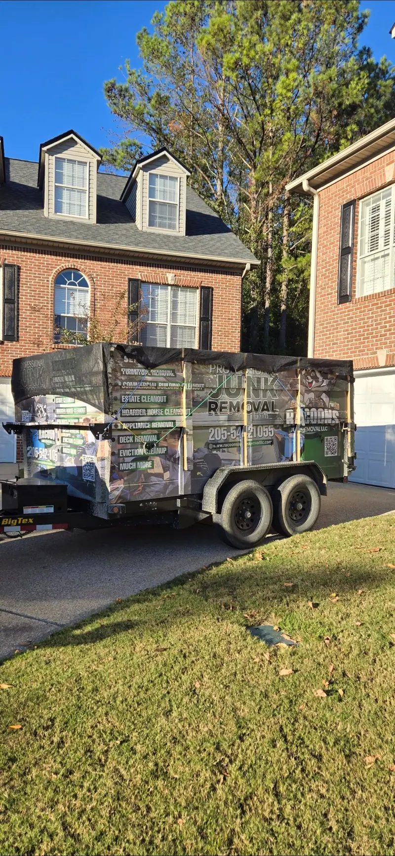 The Racoons Junk Removal dump trailer, wrapped with the racoon logo, the phone number and a list of services, parked in a Birmingham-area driveway